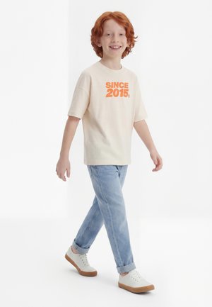 Mosolygó, vörös hajú gyermek sétál, krémszínű pólót visel narancssárga "SINCE 2015" felirattal, világoskék farmert és fehér tornacipőt barna talppal.