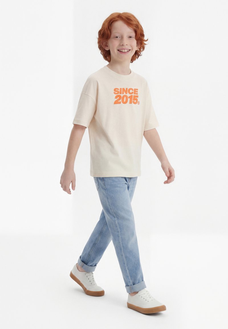 Χαμογελαστό παιδί με κόκκινα μαλλιά που περπατά, φοράει κρεμ μπλουζάκι με τη φράση "ΑΠΟ ΤΟ 2015" σε πορτοκαλί, ανοιχτό μπλε τζιν και λευκά αθλητικά παπούτσια με καφέ σόλες.