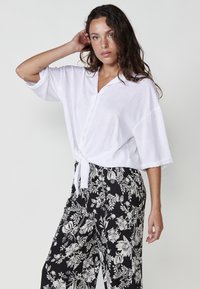 Camicia bianca con collo a cravatta, vestibilità comoda e maniche corte, abbinata a pantaloni larghi a fantasia floreale nera.