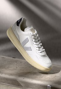 Sneaker bianca in pelle con punta perforata, logo testurizzato argento e suola in gomma beige. Presenta un linguetta sul tallone di colore blu scuro e lacci.