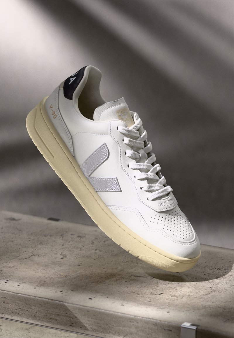 Sneaker bianca in pelle con punta perforata, logo testurizzato argento e suola in gomma beige. Presenta un linguetta sul tallone di colore blu scuro e lacci.