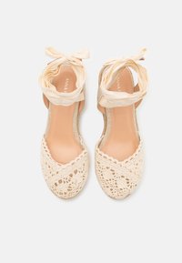 Beige kanten espadrilles met een geweven haakwerkdesign, open toe en stoffen enkelbandjes. De zool is natuurkleurig en plat.