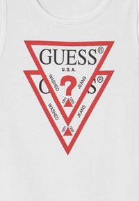 Biały top na ramiączkach z czerwonym trójkątnym grafikiem z napisem "GUESS U.S.A." oraz słowami "WASHED" i "JEANS" na czarno, a także z pytajnikami i cyframi.