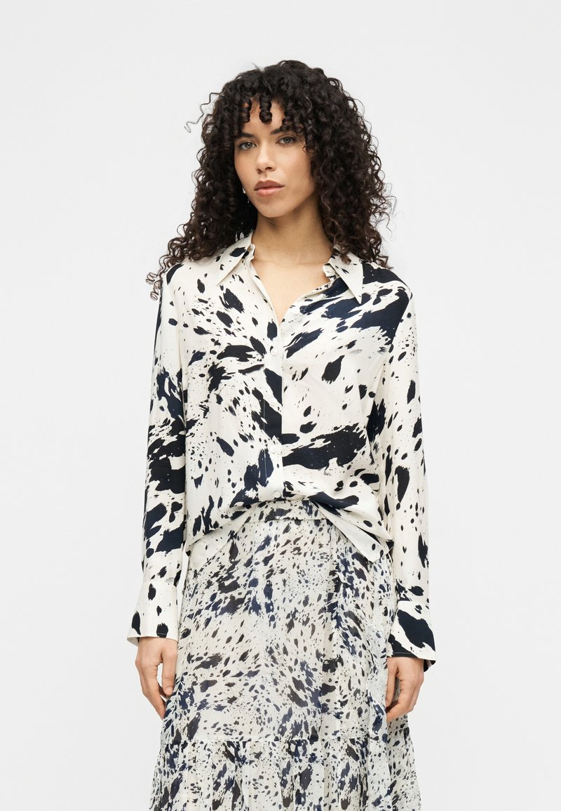 Πουκάμισο με μακριά μανίκια σε εκρού χρώμα με μαύρα σχέδια που μοιάζουν με πιτσιλιές, σε συνδυασμό με φούστα πολλαπλών επιπέδων στην ίδια απόχρωση. Και τα δύο είναι κατασκευασμένα από ελαφρύ, ρέον ύφασμα.