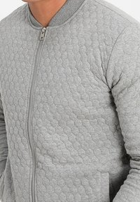 Giacca bomber grigia trapuntata con chiusura a zip, caratterizzata da un motivo esagonale testurizzato e colletto, polsini e orlo a coste.