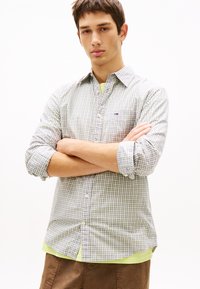 Νεαρός άνδρας με κοντά καστανά μαλλιά στέκεται με σταυρωμένα τα χέρια, φορώντας ανοιχτό γκρι καρό πουκάμισο πάνω από ένα λαχανί πράσινο T-shirt και καφέ παντελόνι.