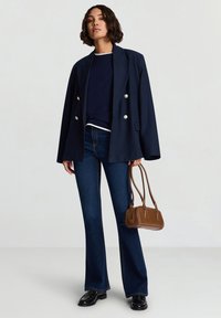 Marineblazer mit doppelten Knöpfen, kombiniert mit einem dunklen Oberteil, ausgestellten blauen Jeans und einer kleinen braunen Handtasche. Schwarze Schuhe runden das Outfit ab.