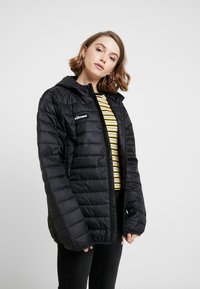 Doudoune noire avec fermeture éclair et capuche. Présente un design matelassé et un logo sur la poitrine gauche. Portée par-dessus une chemise rayée.