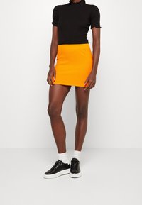 Černý žebrovaný top s krátkým rukávem spárovaný s texturovanou oranžovou mini sukní. Model má na sobě černé tenisky s bílou podrážkou a bílé kotníkové ponožky.