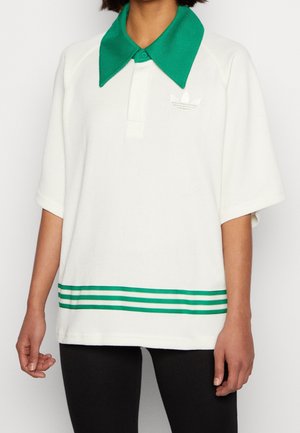 Kobieta w białej krótkim rękawem dzianinowej polówce z zielonym kołnierzykiem i trzema zielonymi paskami przy dolnym brzegu, w połączeniu z czarnymi legginsami.