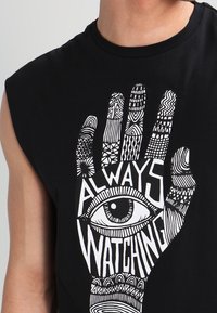 Czarna bezrękawnik z białym nadrukiem przedstawiającym dłoń i oko z misternymi wzorami oraz tekstem "ZAWSZE PATRZY".