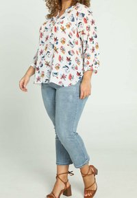 Blouse blanche avec imprimés colorés d'animaux et de fleurs, manches trois-quarts, associée à un jean bleu clair retroussé et des sandales marron à talons.