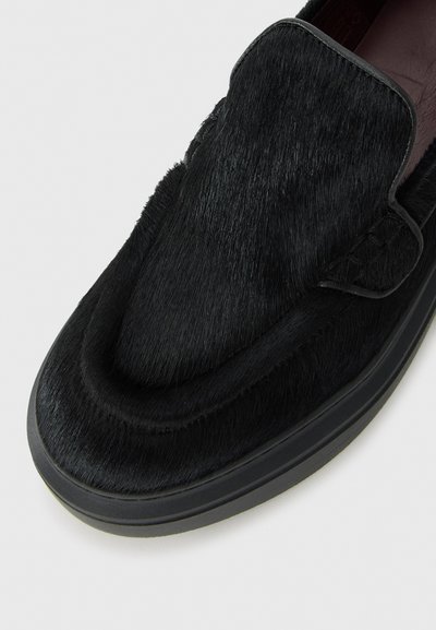 Mocassino in pelle nera con superficie texturizzata e suola in gomma spessa, inclinato per mostrare la parte anteriore e il lato.