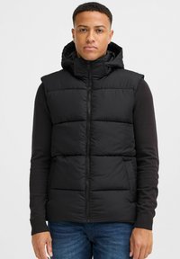 Schwarze Steppweste mit Kapuze, mit Reißverschluss, gestepptem Design und seitlichen Taschen. Trägt man über einem dunklen Langarm-Oberteil.