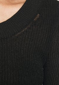 Gros plan d'un pull en maille côtelée noire avec une petite déchirure horizontale près de l'encolure sur l'épaule droite.