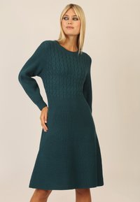 Teal knælængde kjole med kabelstrik tekstur på overdelen og ribbet stoff på ærmerne og nederdelen, med en klassisk rund halsudskæring.