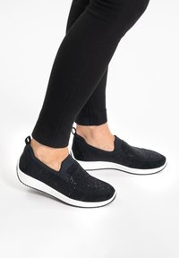 Schwarze Slipper mit einem strukturierten Obermaterial, das ein strickartiges Design aufweist und eine weiße Gummisohle hat. Mit einem Tab an der Ferse akzentuiert.