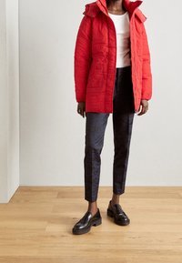 Rode gewatteerde pufferjack met een capuchon, rits vooraan en zijzakken, gecombineerd met donkere met een patroon versierde broeken en zwarte instappers.