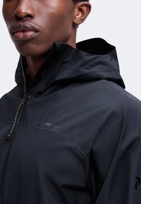 Gros plan d'une personne portant une veste à capuche noire avec une fermeture éclair à l'avant et une discrète marque sur la poitrine et la manche.