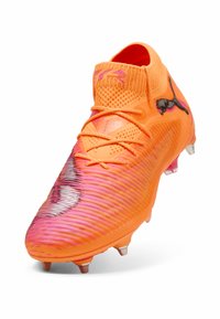 Scarpino da calcio arancione brillante con un design testurizzato a gradiente che sfuma nel rosa. Presenta strisce diagonali, un collare alto e dettagli metallici.