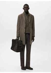 Abrigo de lana marrón sobre un suéter de punto oscuro, combinado con pantalones a juego y zapatos de ante, sosteniendo un bolso de ante oscuro, sobre un fondo blanco.