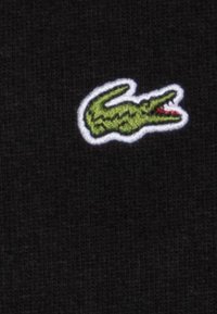 Juodas audinys su raukšlėta tekstūra, kuriame išsiuvinėtas žalias krokodilo logotipas, aprašytas balta spalva. Dizainas yra centrinėje pozicijoje.