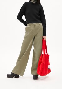 Pull noir côtelé, pantalon large kaki, sabots noirs, et un sac fourre-tout rouge texturé avec un logo sur le côté.