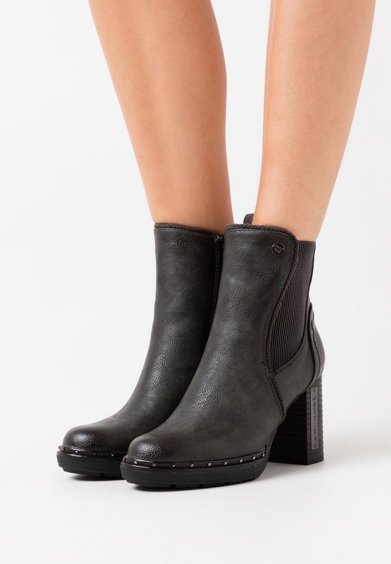 Bottines en cuir noir à talons hauts avec panneaux élastiques sur les côtés, bordure cloutée et talons blocs, portées sur jambes nues sur fond blanc.