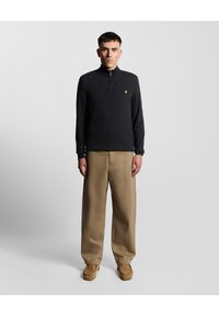 Svart zip-up tröja med hög krage, prydd med en liten gul logotyp. I kombination med beiga vida byxor och bruna skor.