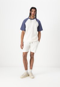 Camiseta de manga corta en color crema con mangas azul marino, combinada con pantalones cortos ligeros de textura. El calzado consiste en unas zapatillas beige con calcetines blancos.