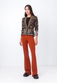 Giacca ricamata multicolore con base nera, motivi floreali e dettagli dorati, abbinata a pantaloni svasati color ruggine e scarpe con plateau nere.