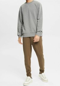 Felpa grigia con maniche lunghe e scollo rotondo, abbinata a pantaloni jogger marroni in pile. Le scarpe sono bianche con dettagli neri.