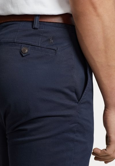 Man die marineblauwe broek draagt met een knoopsluiting op de achterzak, een bruine leren riem en een wit overhemd dat in de broek is gestopt, gezien van de taille naar beneden.