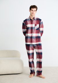 Hombre de pie descalzo sobre una alfombra, vistiendo un conjunto de pijama de cuadros en rojo, blanco y marino con mangas largas y pantalones a juego, al lado de un sofá curvado beige.