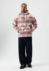 Sweat à capuche en tissu doux avec un motif rouge et beige d'animaux et de cactus ; porté avec un jean foncé et des chaussures noires.