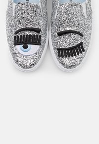 Kimalaiset hopeiset slip-on kengät, joissa on brodeeratut silmämotivit ripseineen, sileä pinta ja valkoinen kumipohja.