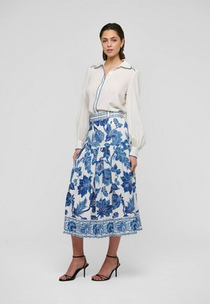 Lange mouwen, crèmekleurige blouse met blauwe biezen, gecombineerd met een zwierige, witte rok met een blauw bloemenpatroon en kanten zoom. Zwarte sandaalhakken met bandjes.