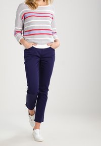 Žena v bílém svetr s červenými a fialovými pruhy, tmavě modrých kalhotách, bílých slip-on botách, s rukama v kapsách, kráčející vpřed.