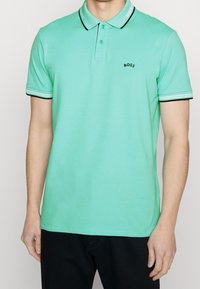 Ljusgrön polo skjorta med svart och vit randig krage och mudd. Har tre knappar vid halsen och en liten svart logotyp på bröstet.