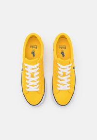 Gula canvas-sneakers med vita snören, blå accenter och en logotyp på tungan. Designen har en lågtopp-silhuett och gummisula.