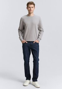 Grijs langarmshirt met een ronde hals, gecombineerd met donkerblauwe spijkerbroek en witte sneakers. Model staat met handen in de zakken.