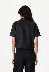 Camisa preta de mangas curtas com colarinho, corte estruturado, textura suave e design cropped. A parte de trás é lisa, sem detalhes visíveis.