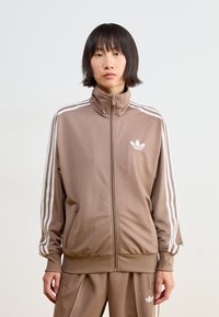 Braune zip-up Trainingsjacke mit weißen Streifen, hohem Kragen und Logo auf der Brust. Weicher Stoff und lockere Passform. Passende Hose sichtbar.