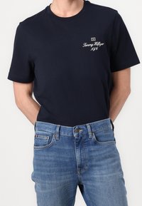Hombre vistiendo una camiseta navy blue de Tommy Hilfiger con el logo en el pecho y jeans de mezclilla azul, de pie con las manos detrás de la espalda.