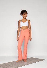 Rózsaszín, sztreccs anyagból készült bővülő nadrág, fehér sportmelltartóval párosítva. A modell egy jóga matracon áll, mosolyogva, jól megvilágított környezetben.