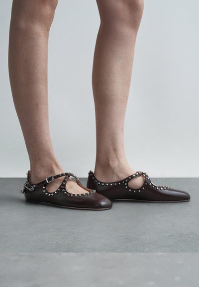 Bailarinas de cuero marrón con punta afilada, con laterales recortados, acentos plateados de tachuelas y detalles de hebilla en la correa. Textura suave.