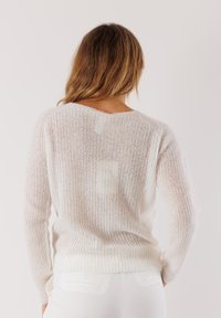 Donna con capelli lunghi che indossa un maglione bianco trasparente e pantaloni bianchi, in piedi contro uno sfondo chiaro, vista da dietro.