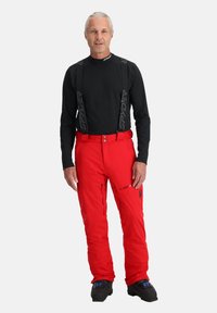 Rote wasserdichte Skihosen mit verstellbaren Trägern, Reißverschlusstaschen und schwarzen Akzenten. Getragen mit einem schwarzen Langarmshirt.