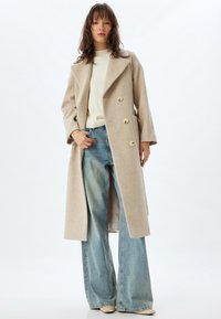 Abrigo de lana beige con cuello con muescas, botones de doble botonadura y bolsillos laterales, combinado con jeans de pierna ancha de color azul claro.