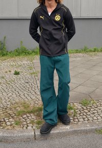 Persona che indossa una giacca zip-up nera del Borussia Dortmund, pantaloni larghi turchesi e scarpe nere, in piedi su un marciapiede.